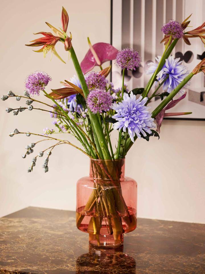 Bloemenboeket in een roze glazen vaas met paarse alliums, blauwe chrysanten en diverse andere bloemen, op een marmeren oppervlak.