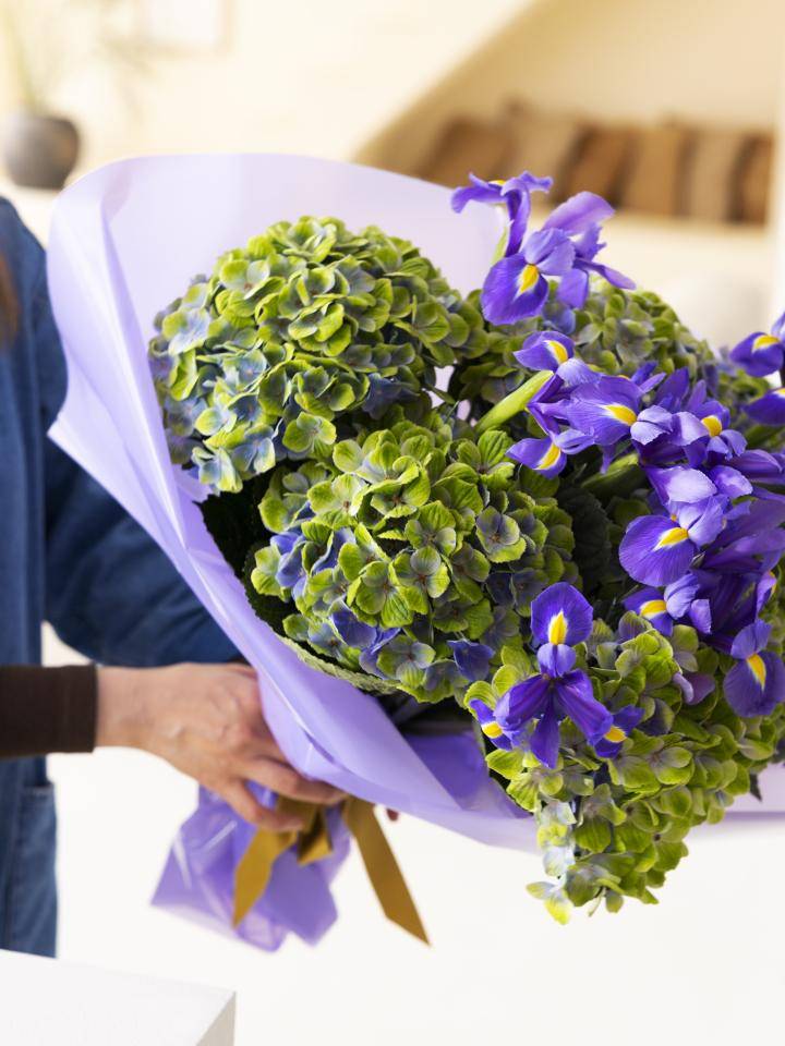 Persoon met een boeket van groene en paarse hortensia's en paarse irissen, gewikkeld in lichtpaars papier met een bruine lint.