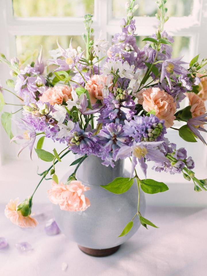 Vaas met diverse bloemen, waaronder lichtpaarse en perzikkleurige bloemen, op een lichtgekleurde ondergrond met paarse kristallen, bij een raam met natuurlijk licht.