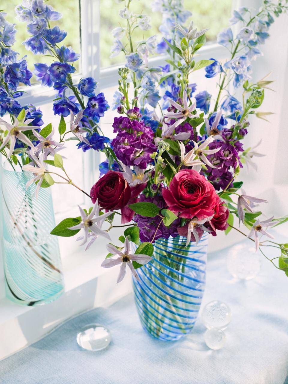 Boeket bloemen in een blauw-wit gestreepte vaas, met rode rozen, paarse bloemen en blauwe riddersporen, bij een raam met natuurlijk licht.
