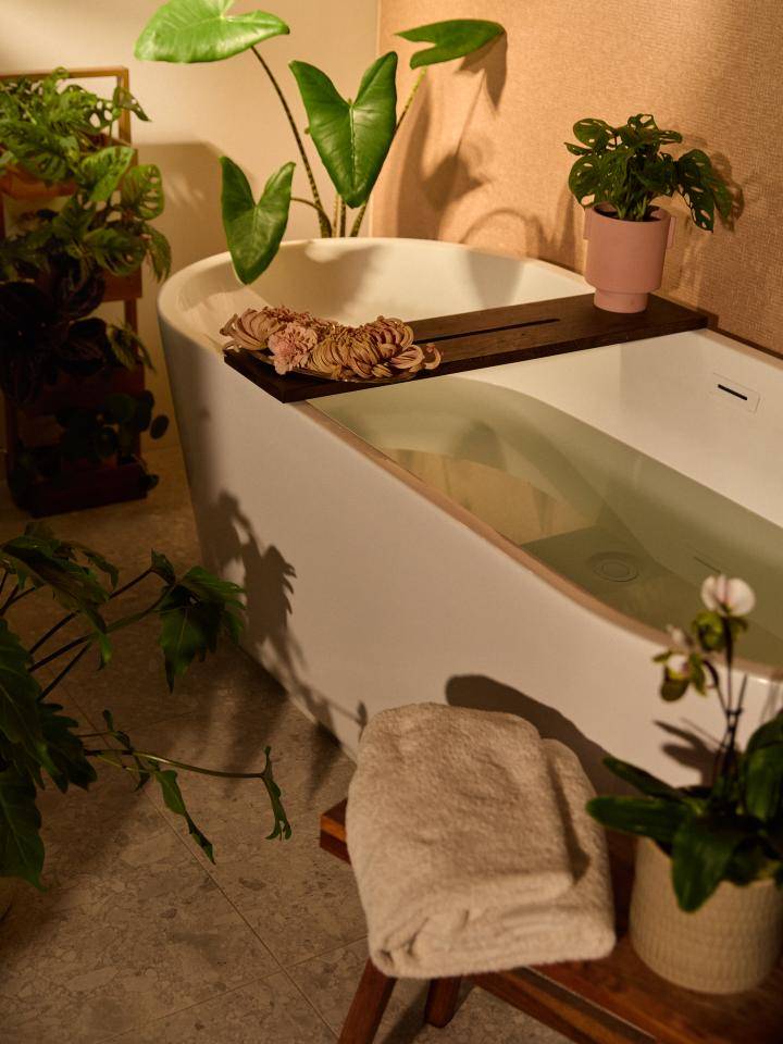 Badewanne umgeben von grünen Pflanzen, mit einem Handtuch auf einem Holzschemel.
