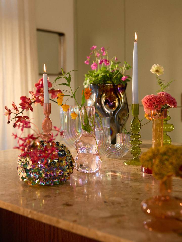 Verschiedene dekorative Gegenstände auf einer Marmorarbeitsplatte, darunter Vasen mit Blumen und zwei Kerzen in rosa und grünen Haltern, mit einem Hintergrund aus Vorhängen und einem Spiegel oder Bilderrahmen.