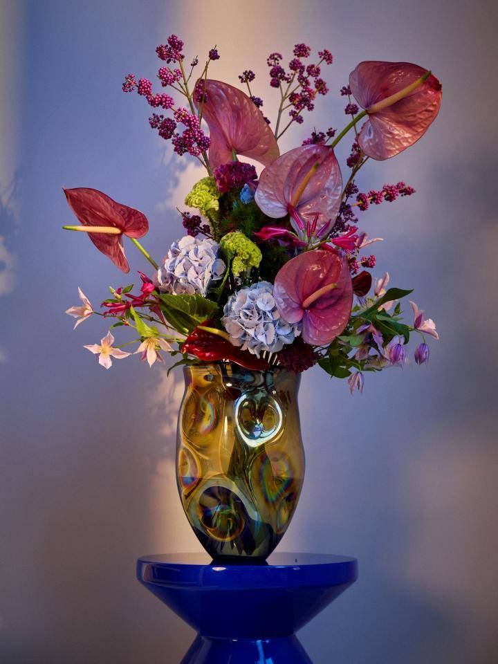 Bloemstuk in een vaas op een blauwe sokkel, met roze anthuriums, paarse hortensia's en ander groen.