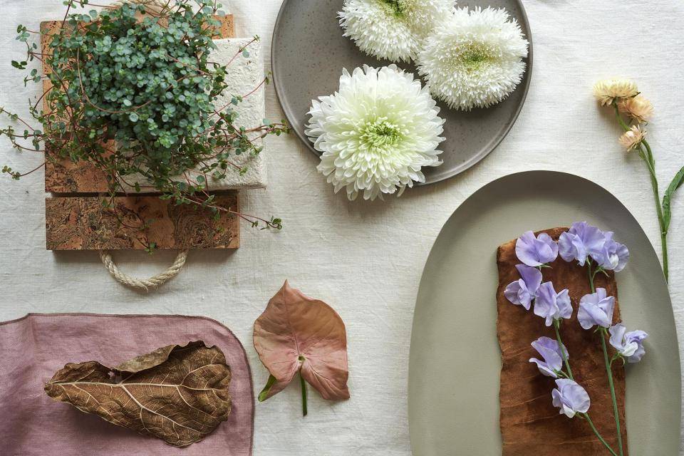 Arrangement von Pflanzen und Blumen auf einem hellen Stoff, mit einer Topfpflanze, Chrysanthemen und getrockneten Blättern.