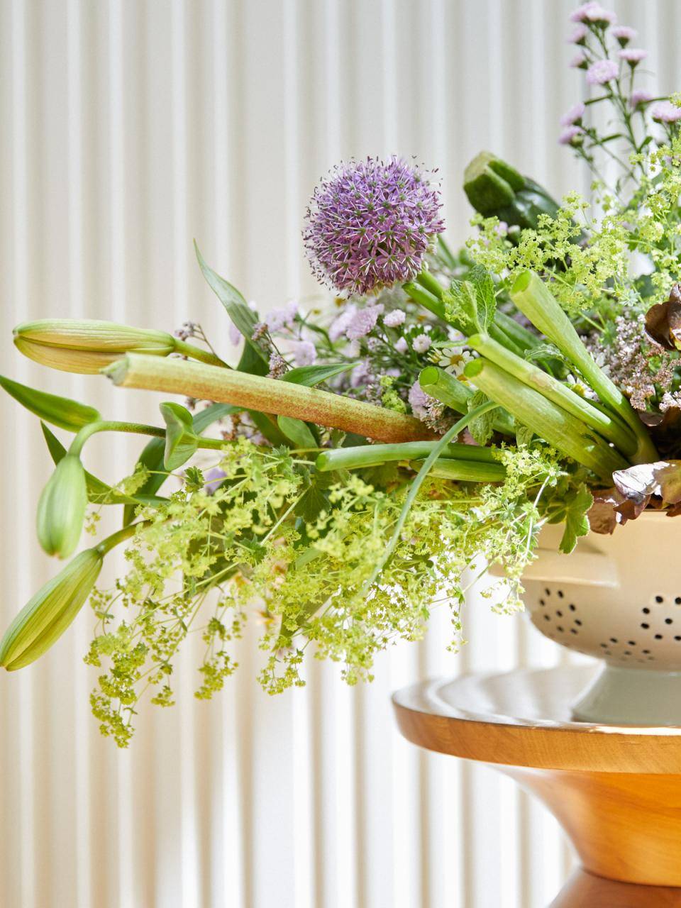 Bloemstuk in een wit vergiet op houten standaard, met paarse bloem in het midden.