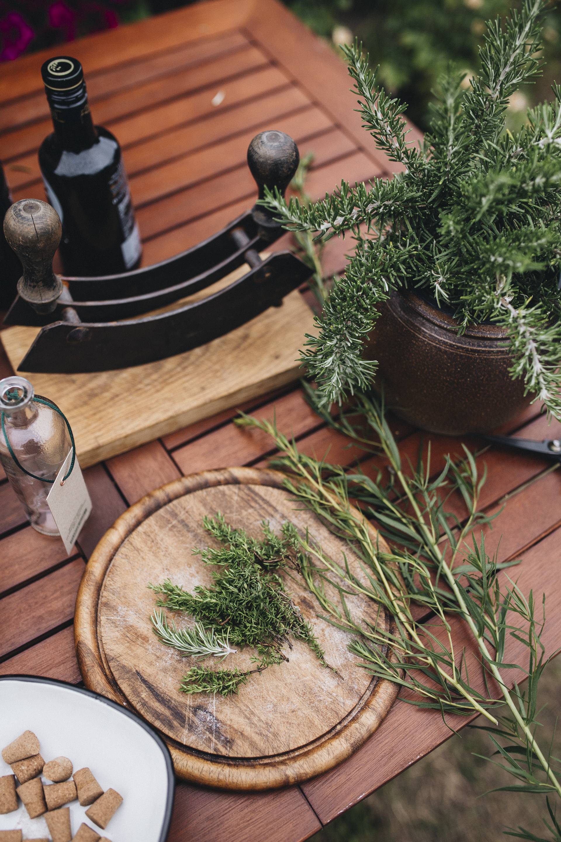 Rozemarijn op een houten snijplank met een witte kom en een bruine pot op een houten tafel.