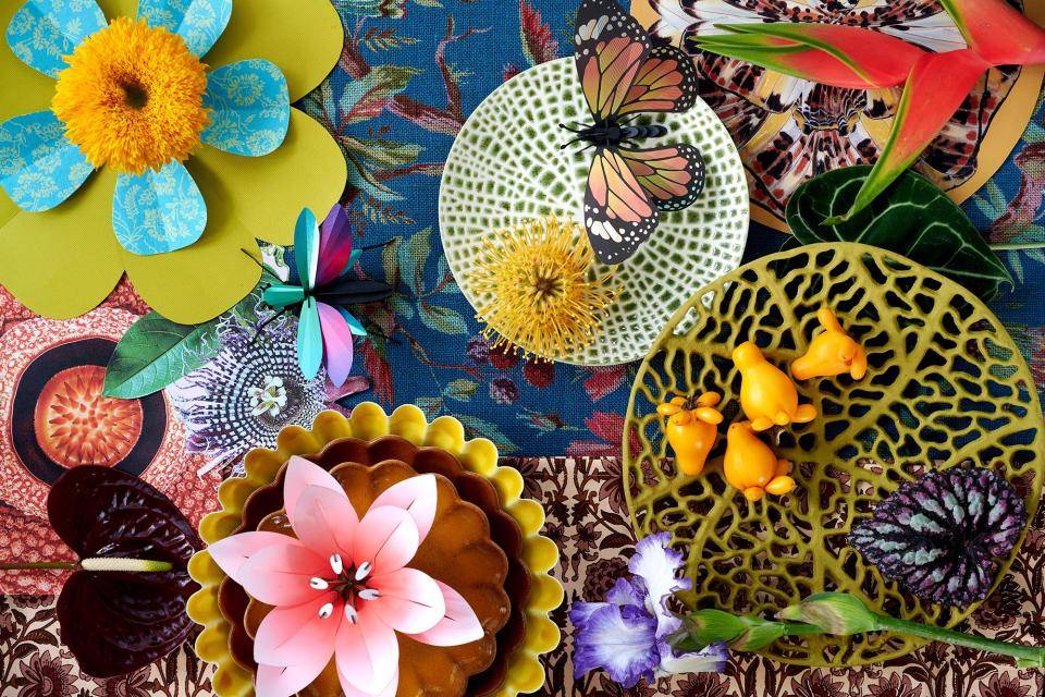 Farbenfrohes Arrangement von Blumen, Blättern, Schmetterlingen und dekorativen Tellern auf gemusterten Stoffen.