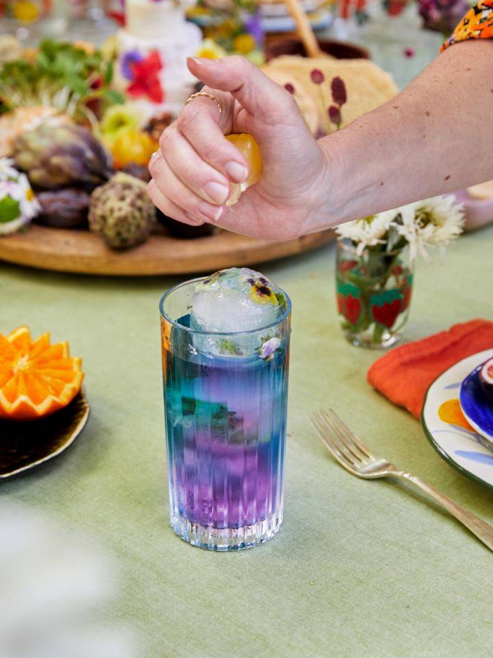 Hand knijpt citroen over kleurrijke mocktail met ijs en bloemen op tafel met eten.