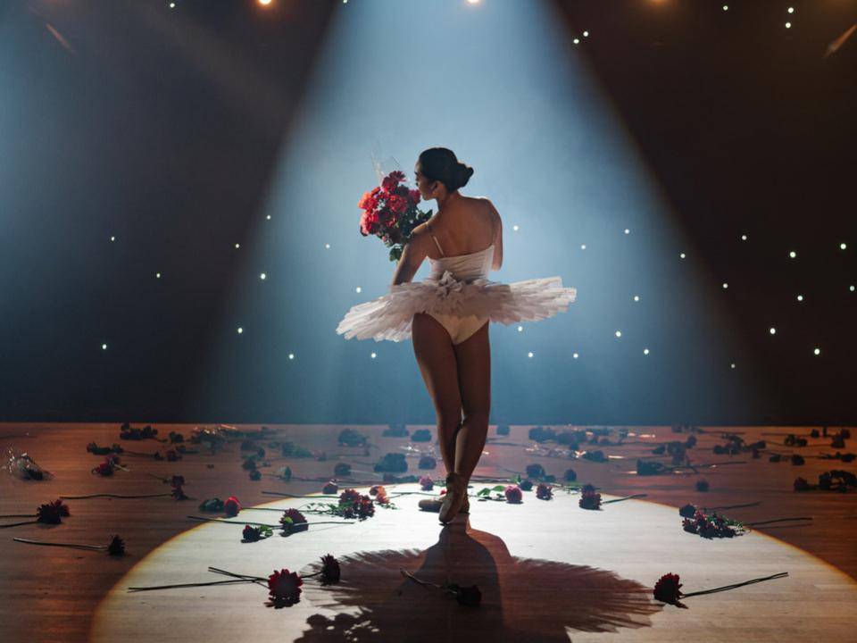 Balletdanseres in witte tutu houdt een boeket rode bloemen vast op een podium, omringd door losse bloemen.
