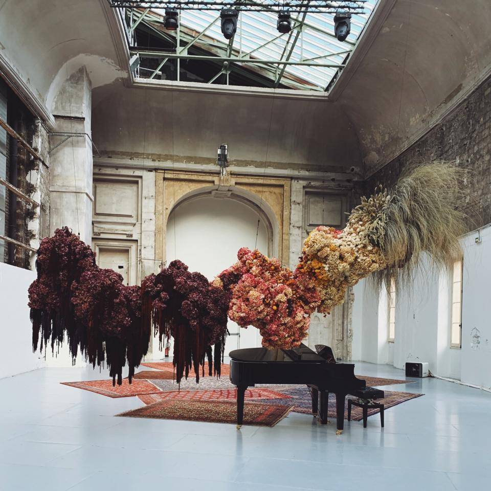 Kunstinstallatie met twee grote, hangende bloemstukken, een vleugel en oosterse tapijten in een ruimte met hoog plafond.