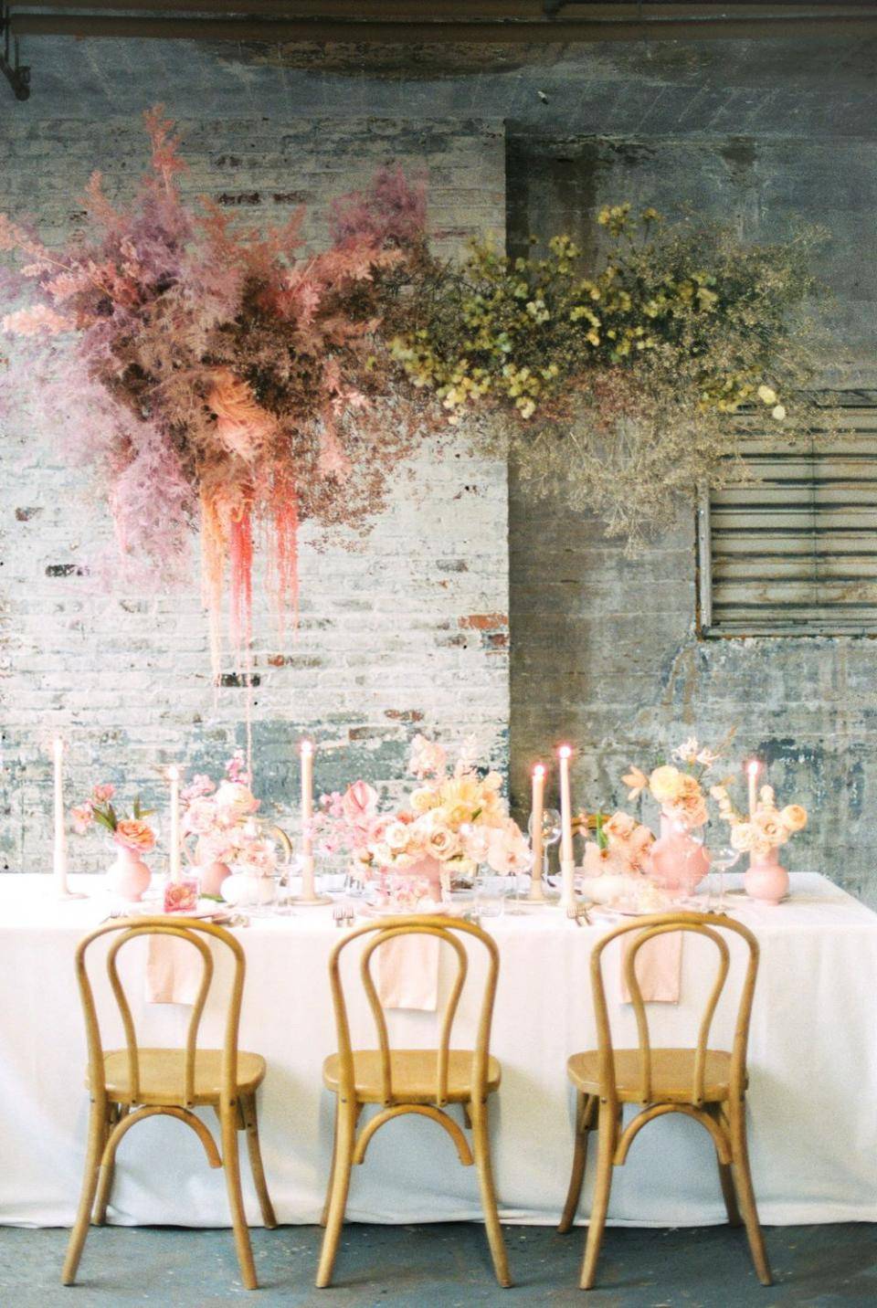 Eettafel met witte tafelkleed en bloemenarrangementen, tegen een rustieke bakstenen muur.
