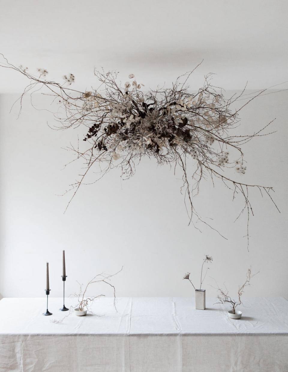 Witte tafel met gedroogde takken en bloemen, twee kandelaars en een vaas, tegen een witte achtergrond.