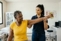 Zorgprofessional begeleidt oudere vrouw bij arm- en schouderoefeningen in een behandelruimte van een gezondheidscentrum.