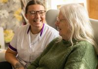 Verzorgende en oudere vrouw lachen samen tijdens een gesprek in een huiselijke omgeving.